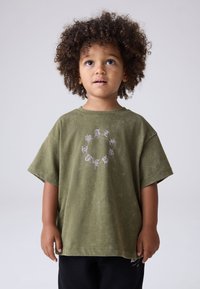 Ungt barn med lockigt hår som b�är en oversize olivgrön t-shirt och svarta byxor, tittar lite uppåt mot en enfärgad bakgrund.