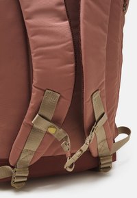 Pink backpack with padded fabric, adjustable tan straps, and gold hardware detailing. Features a textured exterior and reinforced stitching.