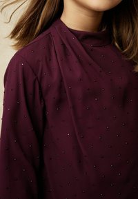 Bordeauxfarbene Bluse mit hohem Ausschnitt, die ein gerafftes Design aufweist und mit kleinen, glänzenden schwarzen und lila Perlen verziert ist.