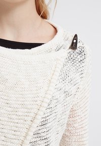 Pull en tricot blanc avec un décolleté bateau ouvert, présentant un motif texturé et un bouton noir en accent sur une épaule.