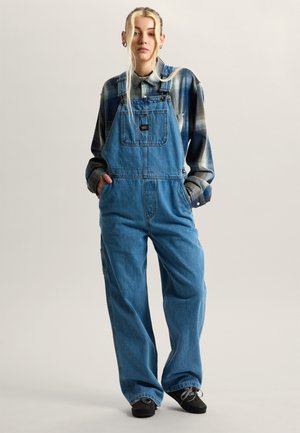 Joven mujer con cabello largo y rubio, vestida con peto de mezclilla azul, camisa a cuadros y zapatillas negras, de pie con las manos en los bolsillos.