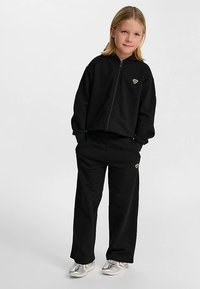 Svart zip-up sweatshirt och matchande byxor, båda tillverkade av mjukt tyg. Har en liten logotyp på bröstet och höften. Enkel, avslappnad passform.