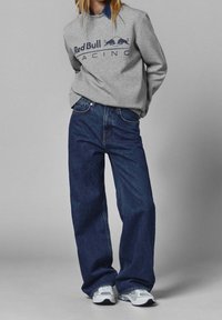 Szara bluza z nadrukiem „Red Bull Racing”, zestawiona z lu�źnymi, ciemnoniebieskimi dżinsami oraz jasnoszarymi butami sportowymi z białymi akcentami.