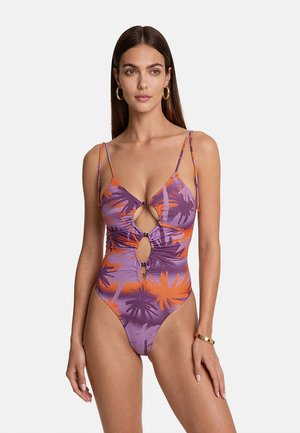 Donna che indossa un costume da bagno viola e arancione con stampa di palme, con ritagli e sottili spalline, accessoriata con orecchini a cerchio dorati e braccialetto.