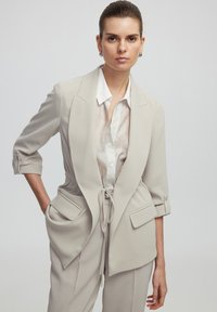 Hellbeige taillierter Blazer mit taillierter Taille, Vordertaschen und hochgekrempelten Ärmeln, über einem weiß gestreiften Hemd kombiniert. Glatte Textur.