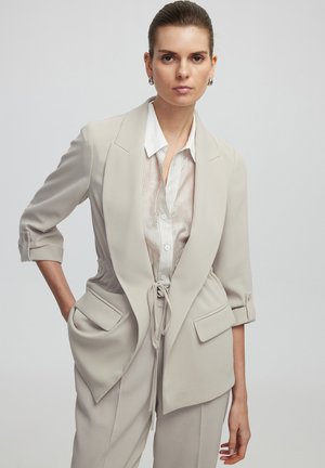 Light beige tailored blazer with a cinched waist, front pockets, and rolled sleeves, layered over a white striped shirt. Smooth texture.