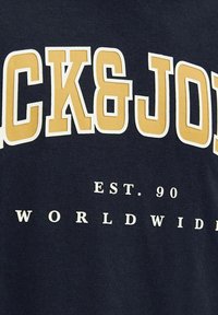 Mornarska majica s hrabrim žutim i bijelim natpisom "CK&JO", s natpisom "EST. 90" i "WORLDWIDE" otisnutim ispod. Pamuk, glatka tekstura.