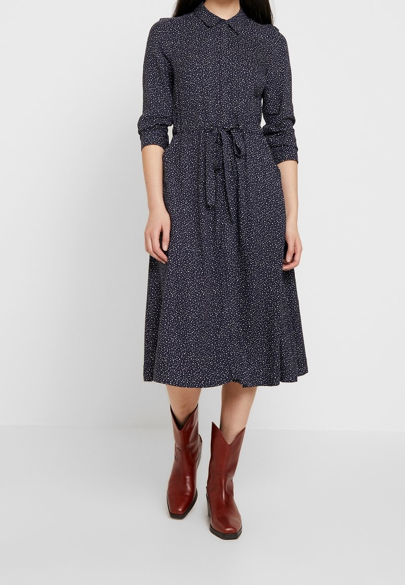 Robe bleu marine à pois blancs, avec col et ceinture à la taille, jupe évasée, portée avec des bottines en cuir marron.