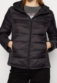 Veste matelassée noire avec fermeture éclair à l'avant, design capitonné, capuche amovible et poches latérales. Texture de tissu lisse et léger.