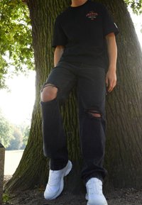 Camiseta negra de gran tamaño con el texto "GRACIOUS", combinada con jeans negros desgastados con rodillas rasgadas y zapatillas deportivas blancas.