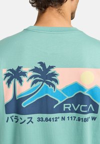 Mintgroene t-shirt met korte mouwen, versierd met een kleurrafische afbeelding van palmbomen, bergen en de zon, met de tekst "RVCA" en coördinaten.