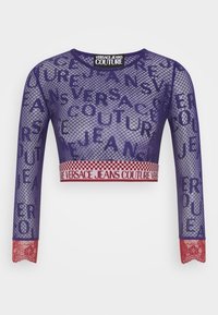 Crop top in rete viola con maniche lunghe, che mostra "VERSACE JEANS COUTURE" in diverse grandezze. Dettagli in pizzo rosso ai polsini. Orlo con banda elastica.