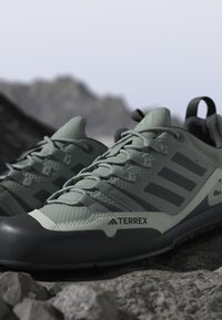 Πράσινα και μαύρα παπούτσια τρεξίματος Adidas Terrex για μονοπάτια τοποθετημένα σε πετρώδη περιοχή με θολό ορεινό φόντο.