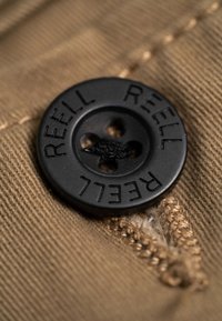 Bouton noir à quatre trous avec l'inscription "REELL" en relief, fixé sur un tissu texturé beige avec des coutures visibles et une petite zone usée.