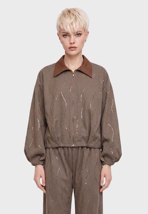 Jeune personne aux cheveux blonds courts porte une veste marron zippée et un pantalon assorti avec des motifs subtils en sequins ondulés.