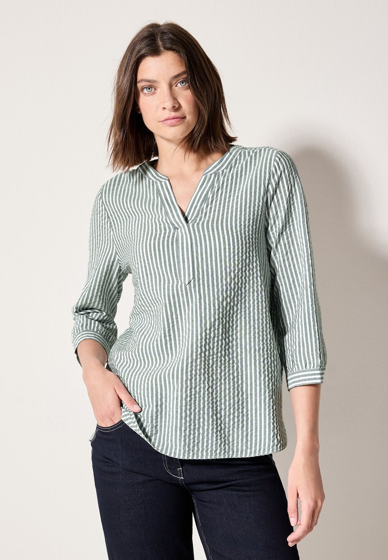 Maglietta a righe verdi e bianche con maniche a 3/4, scollatura a V, tessuto strutturato e vestibilità comoda, abbinata a jeans scuri.