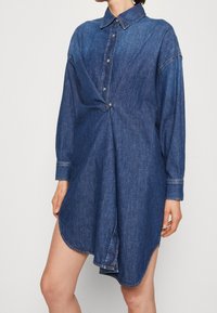 Spijkerjurk in donkerblauw, voorzien van een knoopsluiting aan de voorkant, lange mouwen en een gedraaide taillepartij. Afrondende zoom.