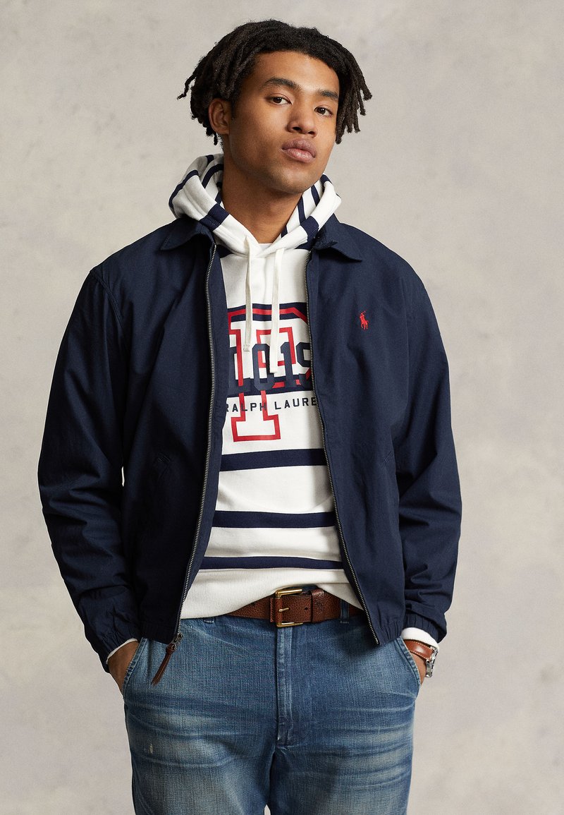 Young man with dark, styled hair wears a navy jacket over a white and navy striped hoodie, paired with light blue jeans, against a gray backdrop.