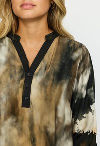 Krageblus med knappar, med ett svart och brunt tie-dye-mönster. Tillverkad av ett slätt tyg med en lätt glans och långa ärmar.