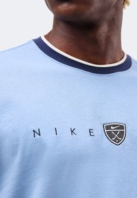 Κοντινό πλάνο ενός ατόμου που φοράει ανοιχτό μπλε πουκάμισο Nike με λευκή και σκούρα μπλε επένδυση στον γιακά και μικρό έμβλημα Nike στο στήθος.