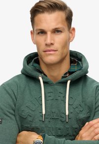 Sudadera verde con un logo texturizado, cordones y una camisa de cuadros en capas debajo. El modelo tiene una expresión neutral y los brazos cruzados.