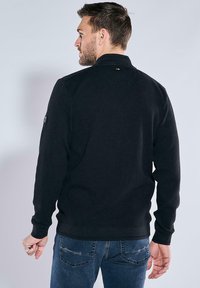 Black textured pullover with a stand-up collar, featuring a stitched pattern and a logo patch on the left sleeve, worn with blue jeans.