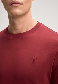 Roter Sweater mit rundem Halsausschnitt, der ein kleines, gesticktes Logo auf der linken Brust und einen glatten, strukturierten Stoff aufweist.