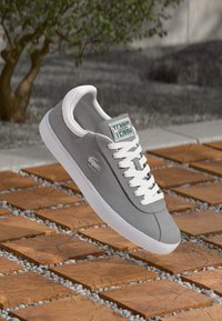 Sivá semišová tenisová obuv s bielymi šnúrkami a podrážkou, logo Lacoste na boku, vznášajúca sa nad drevenými dlaždicami s malými kameňmi vonku.