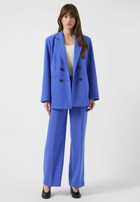 Dobbeltradet blazer og matchende bukser i livlig blå, med svarte knapper og en glatt tekstur. Kombinert med svarte hæler.