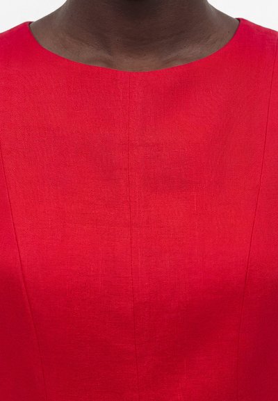 Κοντινό πλάνο ενός ατόμου που φορά ένα κόκκινο φόρεμα με υφή υφάσματος και στρογγυλή λαιμόκοψη, που δείχνει το άνω μέρος του στήθους και του λαιμού.