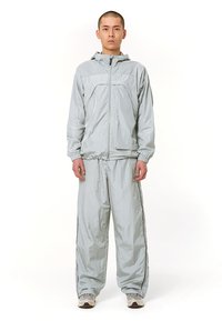 Costum de vânt gri deschis, realizat dintr-un material ușor. Include o jachetă cu fermoar și glugă, cu manșete elastice, asociată cu pantaloni lejeri.