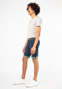 Lichtgrijze t-shirt met korte mouwen, blauwe denim shorts met omslagen en witte sneakers. Model staat zijwaarts tegen een effen achtergrond.