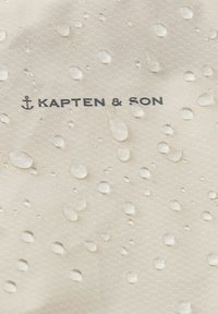 Tessuto beige a motivo esagonale con gocce d'acqua, con il testo "KAPTEN & SON" in grigio scuro. La texture sembra liscia e resistente all'acqua.