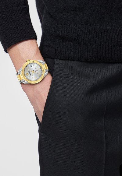 Ezüst és arany színű karóra kerek számlappal, fehér számlappal és dátumkijelzővel. Csuklón viselve, fekete pulóverrel és nadrággal párosítva.