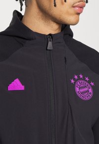 Jeune personne portant un sweat à capuche noir zippé avec un logo Adidas violet et l'emblème du FC Bayern Munich sur la poitrine.