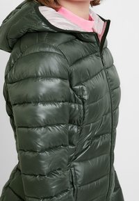 Polstret grøn pufferjakke med en blank overflade, lynlåslukning og hætte. Har vandret syninger og kontrasterende indvendig foring.