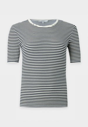 Femme aux longs cheveux blonds portant une chemise rayée noire et blanche, un jean bleu large, des bottes noires, tenant un sac à main noir sur un fond uni.