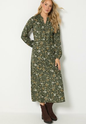 Vrouw draagt een groene midi-jurk met bloemenprint, lange mouwen en een hoge kraag, gecombineerd met donkerbruine enkellaarzen, staand voor een effen achtergrond.