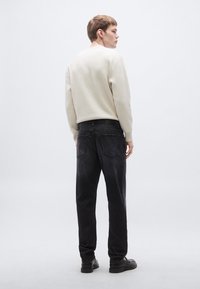 Világos bézs bordázott pulóver, fekete egyenes szárú farmer, és fekete cipő. A nézet hátulról van, bemutatva a ruha szabását és textúráját.