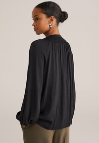 Haját kontyba kötött nő laza, vállain ráncolt, fekete blúzt visel, barna nadrággal párosítva, oldalt állva.