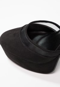 Chaussure à talons hauts en daim noir avec des détails de sangle en maille et un bout pointu fermé sur fond blanc.