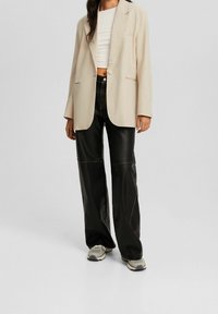 Beżowa, oversize'owa marynarka z długimi rękawami, zestawiona z czarnymi, skórzanymi spodniami o szerokich nogawkach z detalami szwów. Szare sneakersy dopełniają stylizację.