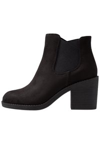 Bottine en suede noir avec un bout arrondi, des panneaux latéraux élastiques et un talon bloc texturé. Texture lisse avec des détails de couture minimaux.