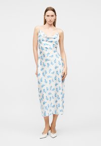 Junge Frau trägt ein weißes Midikleid mit blauem Blumenmuster und weiße spitze High Heels, steht vor einem schlichten hellen Hintergrund.