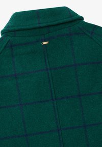 Grønn ullblandingskåpe med en strukturert krage, som har et marineblått rutemønster og et lite gullmerke på baksiden.