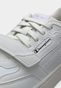 Λευκό αθλητικό παπούτσι με λείο υλικό που μοιάζει με δέρμα, διαθέτει κορδόνια και λουράκι με βέλκρο, διακοσμητικές τρύπες και λογότυπο στο πλάι.