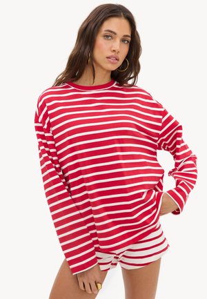 Femme portant un haut à manches longues rouge et blanc à rayures horizontales et un short assorti, main dans la poche et boucles d’oreilles créoles en or.