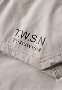 Beiger Stoff mit schwarzem aufgedrucktem Text "T.W.S.N" und einer Zahlenfolge "202315191514" in der Nähe einer genähten Kante.