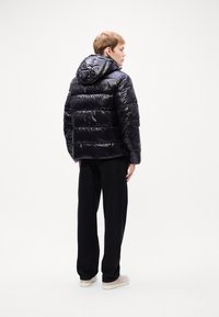 Zwarte pufferjack met capuchon, met een glanzend, gewatteerd oppervlak en horizontale panelen. Draagt over zwarte broeken en lichte sneakers.