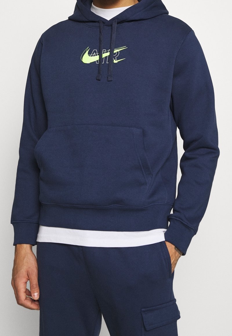 Navy sininen huppari, joka on valmistettu pehmeästä kankaasta ja jossa on edessä kengurupussitasku sekä neonvihreä Nike Air -logo rinnassa.